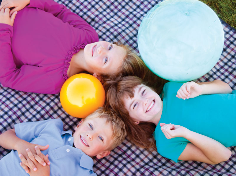 Three kids laying on a blanket outside playing with balls.